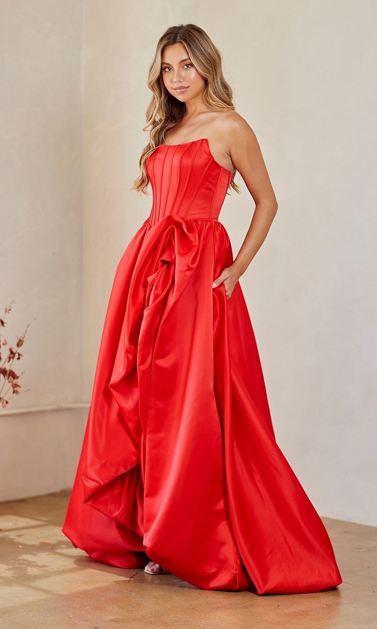 A woman stands indoors wearing the Amelia Couture 2085, a strapless long prom dress featuring a bright red bubble-hem, structured corset bodice, and voluminous skirt.